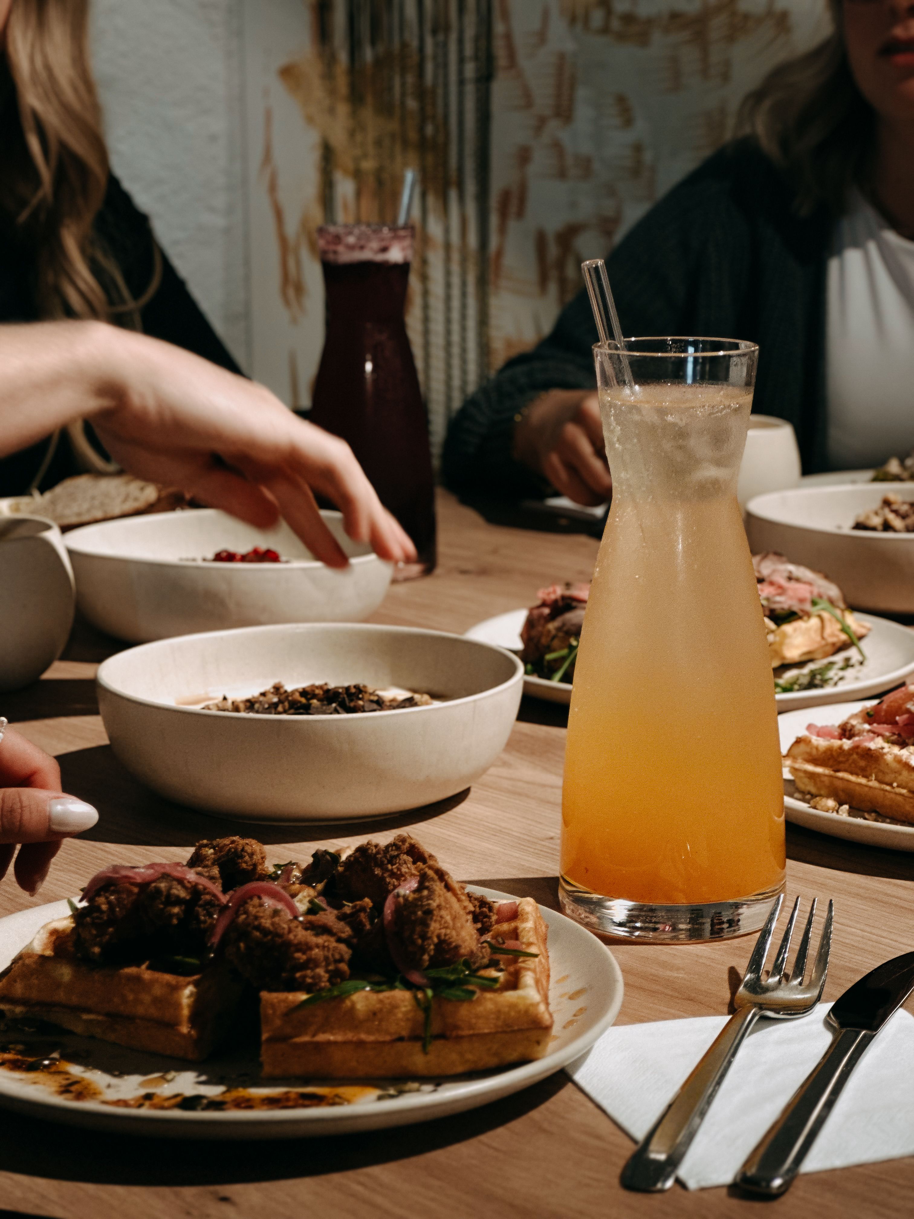 Gedeckter Brunch-Tisch im charmanten Sagmeister Brunch Club mit herzhaften Waffeln, verschiedenen Joghurt-Bowls und hausgemachten Limonaden in Gläsern; mehrere Personen sitzen zusammen und teilen die Speisen in gemütlicher, urbaner Café-Atmosphäre.
